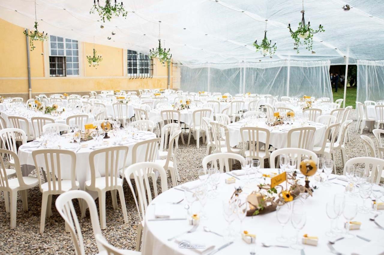 Salle de réception en extérieur sous une tente transparente, avec des tables rondes dressées et décorées.