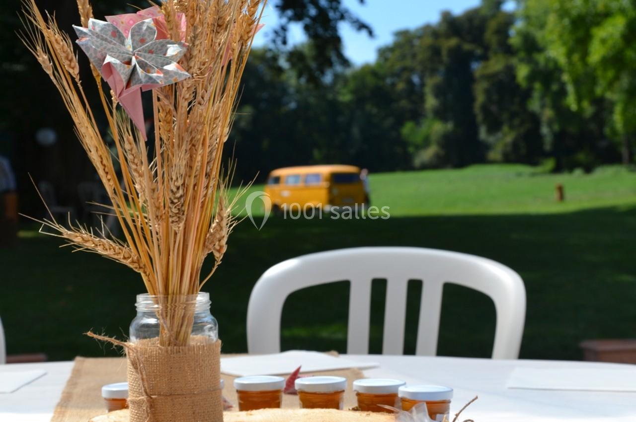 Centre de table champêtre avec épis de blé dans un pot en verre, sur une table extérieure, avec un van jaune en arrière-plan.