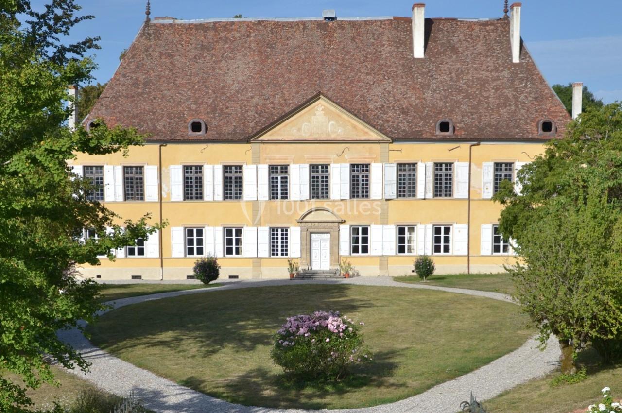 Façade d'un bâtiment historique à deux étages avec un toit en pente, entouré d'un jardin et d'allées gravillonnées.
