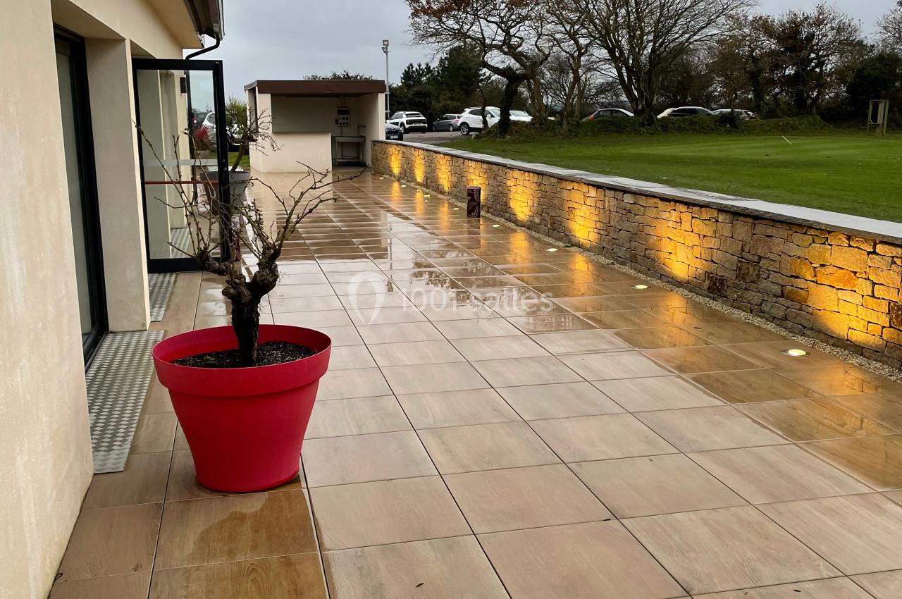 Terrasse en dalles beige mouillée par la pluie, bordée d'un mur éclairé et d'un pot rouge avec un arbuste.
