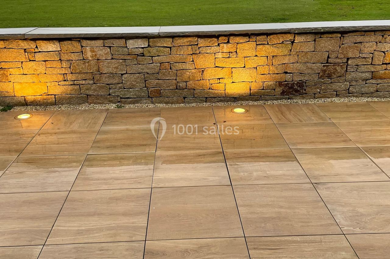 Terrasse en dalles beige clair avec éclairage au sol, mur en pierre et pelouse arborée en arrière-plan.