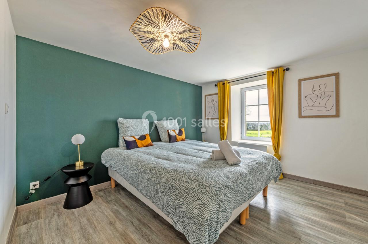 Chambre lumineuse avec lit double, mur vert, rideaux jaunes, parquet clair et lampe design au plafond.
