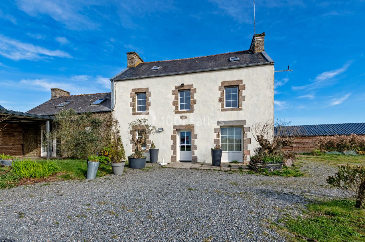 Façade d'une maison en pierre blanche avec des volets marron, entourée d'un jardin et d'une cour gravillonnée.