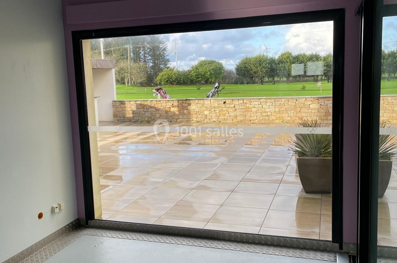 Vue depuis une pièce avec murs violets sur une terrasse carrelée, un jardin et un ciel partiellement nuageux.