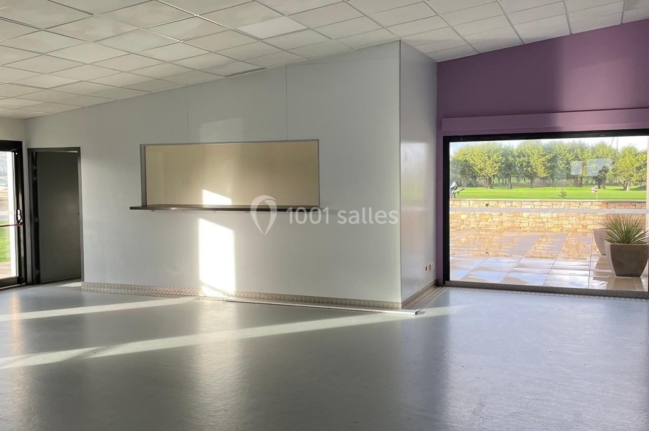 Salle vide avec sol gris, murs blancs, tableau mural, grande fenêtre donnant sur une terrasse et un espace vert.