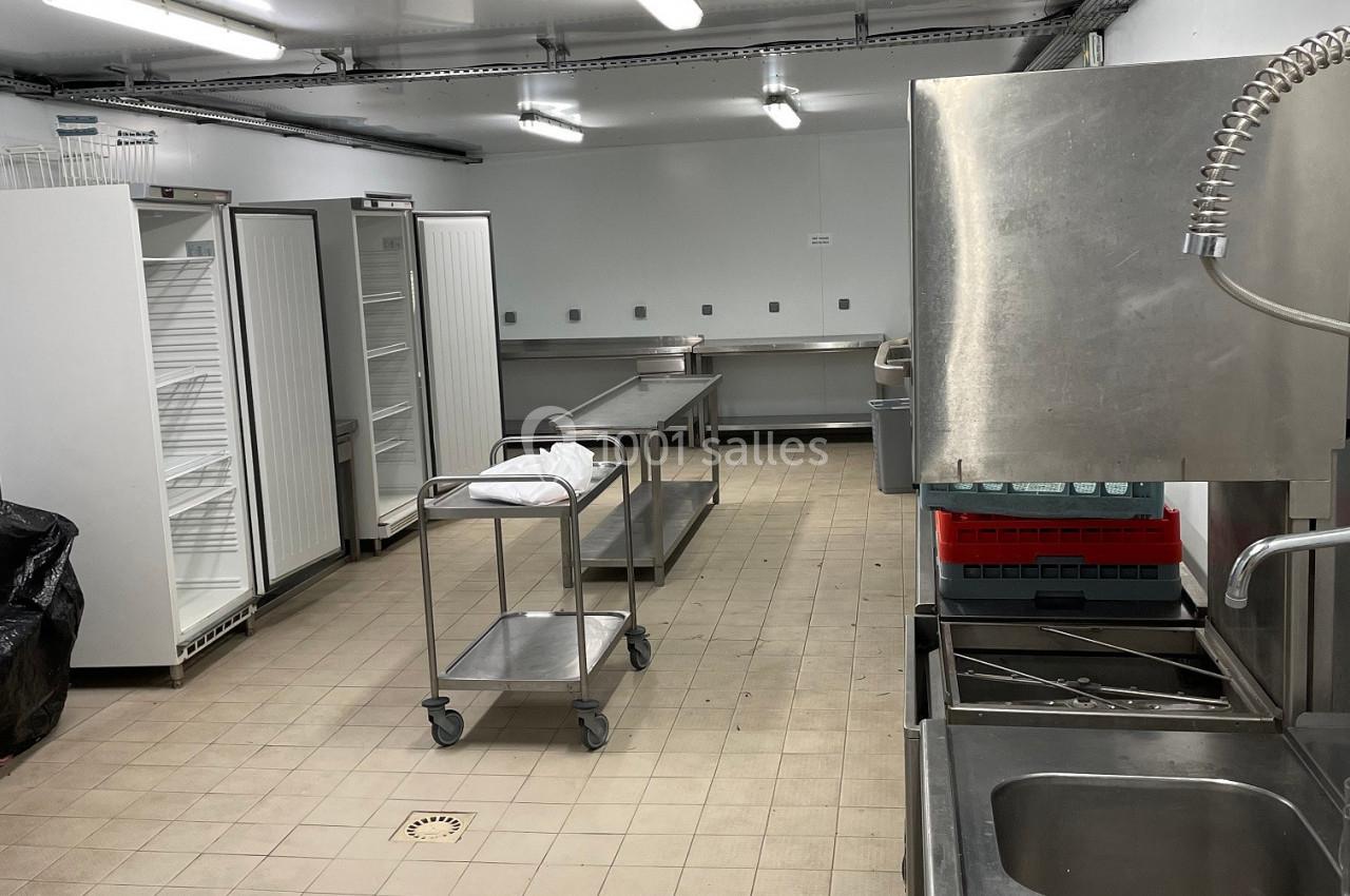 Cuisine professionnelle avec équipements en inox, réfrigérateurs ouverts, plans de travail et sol carrelé beige.