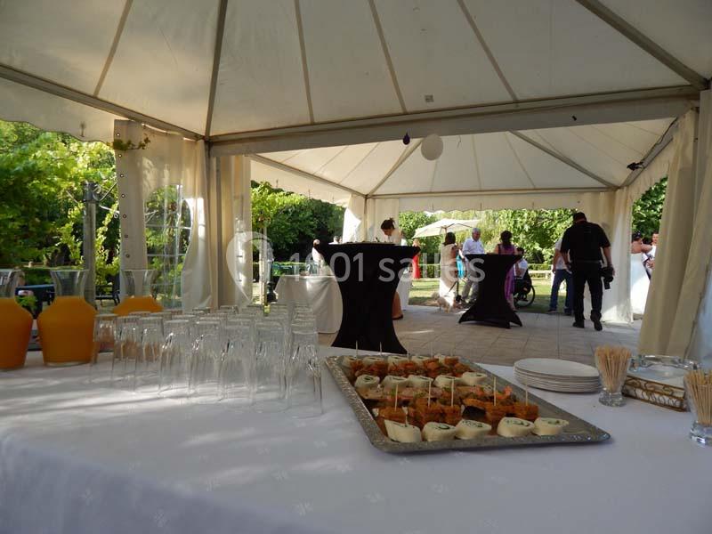 Table dressée avec verrines et boissons sous une tente, en arrière-plan des invités discutent dans un jardin.