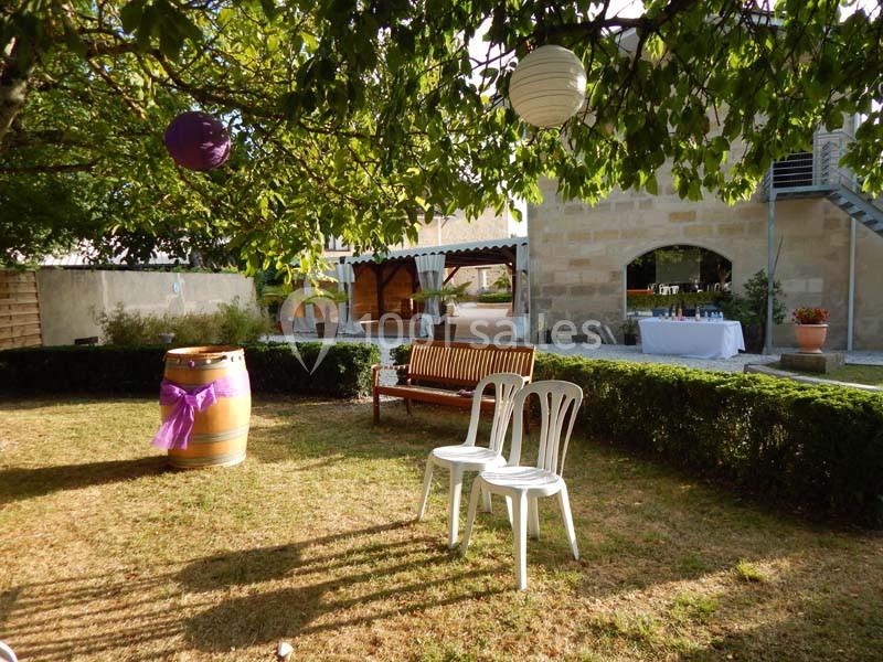 Un jardin avec des chaises blanches, un banc en bois, un tonneau décoré et des lanternes suspendues sous un arbre.