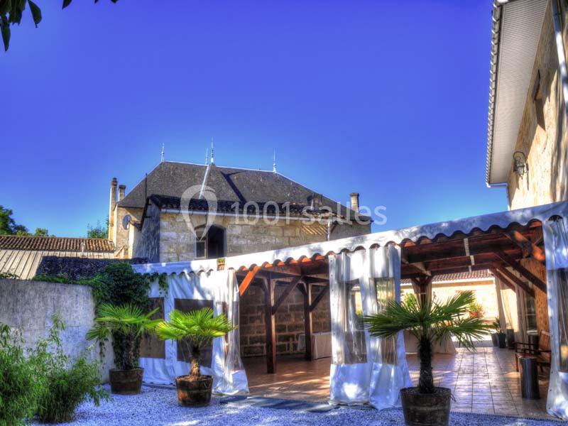 Cour extérieure avec terrasse couverte, palmiers en pots et bâtiment en pierre sous un ciel bleu dégagé.