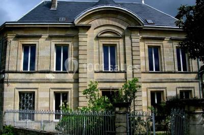 Façade d'une maison en pierre avec toit en ardoise, fenêtres symétriques et jardin clôturé au premier plan.