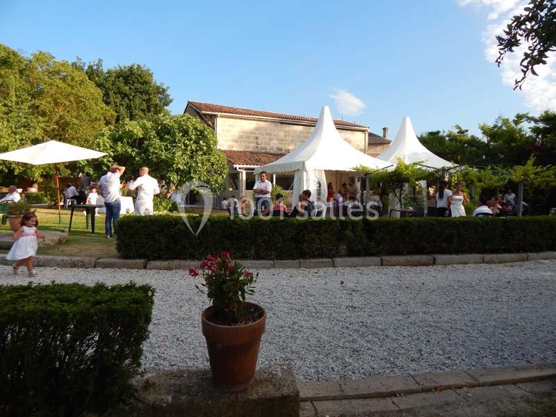 Personnes réunies dans un jardin avec des tentes blanches, entourées de verdure et d'allées gravillonnées.