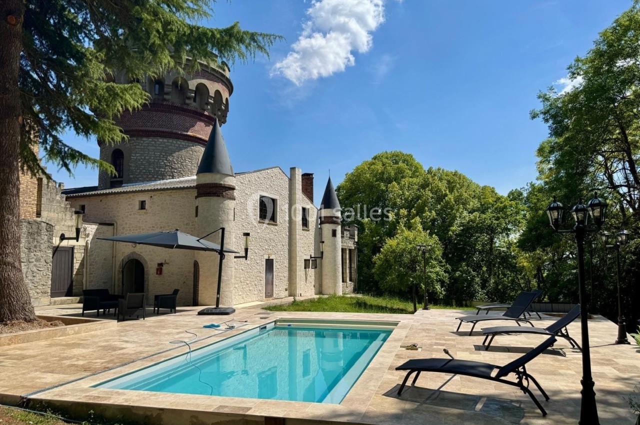 Piscine extérieure entourée de transats et d'un château en pierre avec tourelles, sous un ciel bleu.