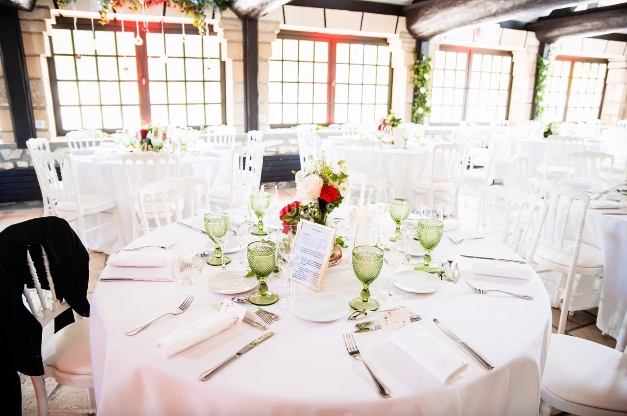 Salle de réception lumineuse avec tables rondes dressées, nappes blanches, verres verts et décoration florale.