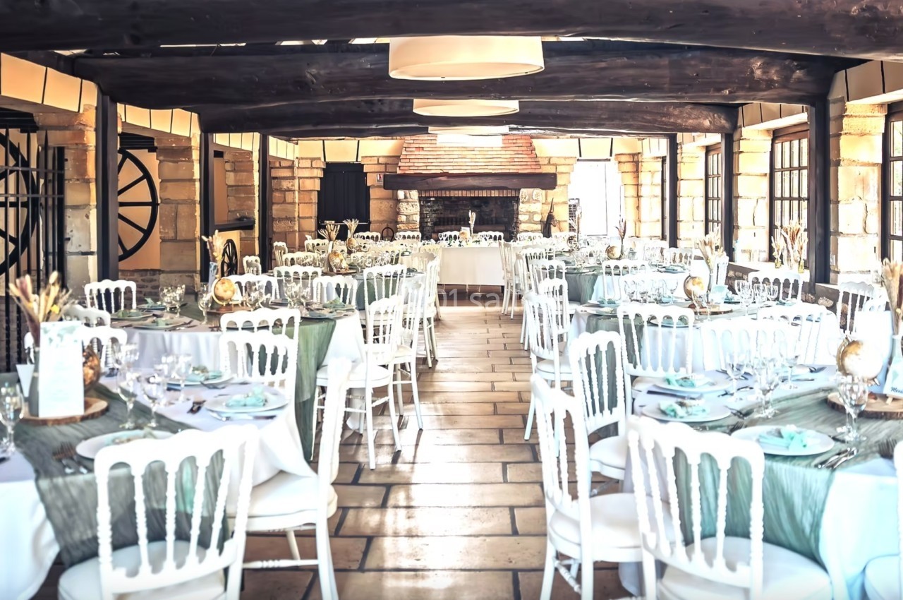 Salle de réception lumineuse avec tables dressées, nappes vertes et blanches, chaises blanches et poutres en bois.