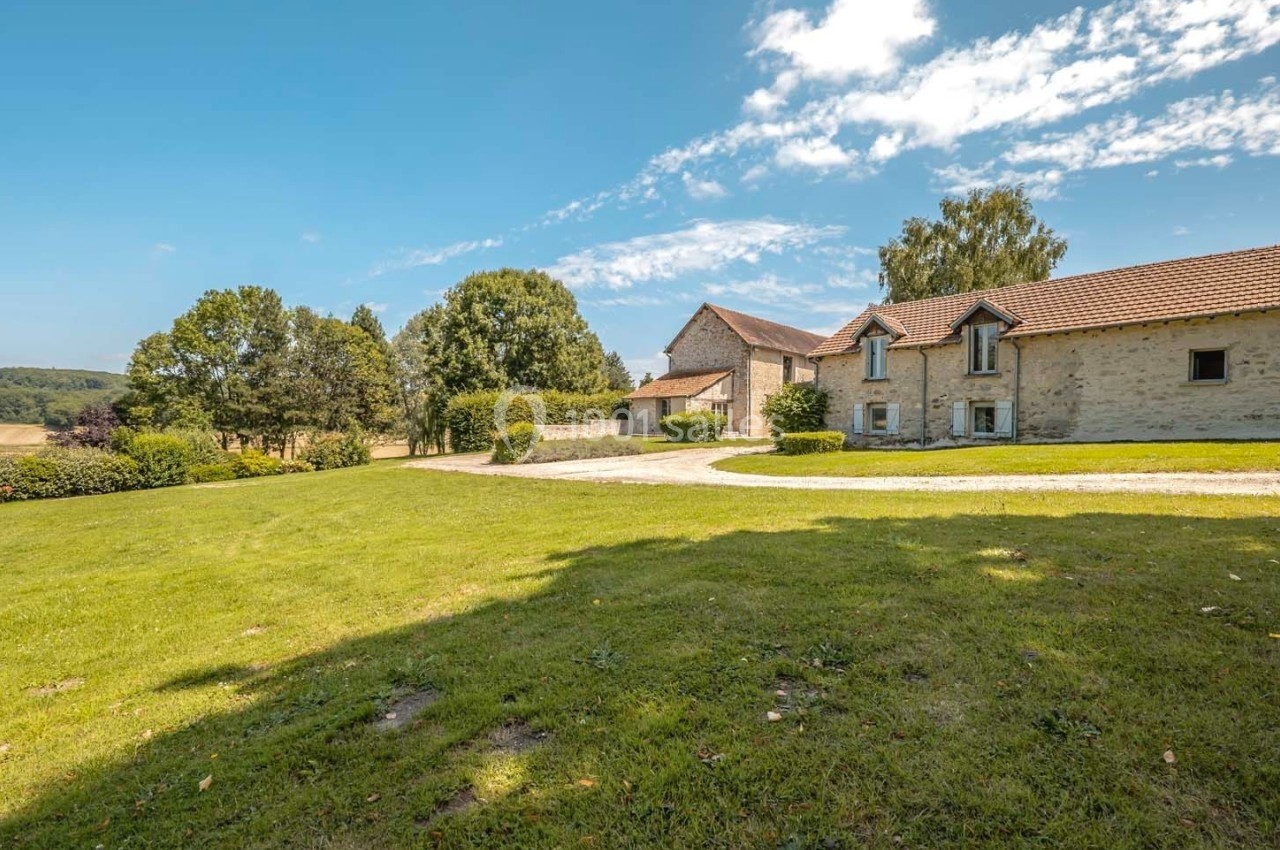 Bâtisse en pierre avec toit en tuiles entourée d'un grand jardin verdoyant sous un ciel bleu.
