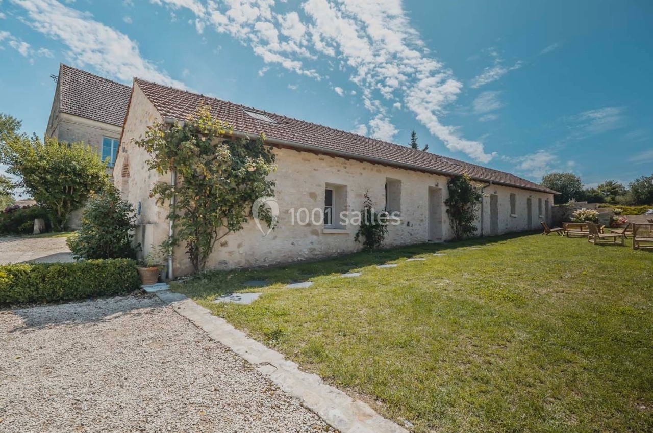 Façade d'une maison en pierre avec toit en tuiles, entourée de verdure et d'un ciel bleu parsemé de nuages.
