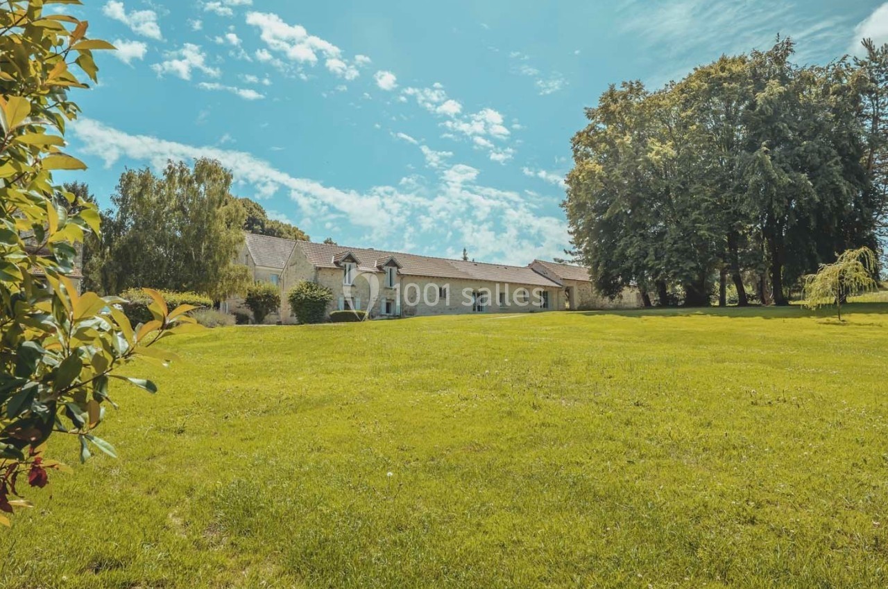 Grande maison en pierre entourée d'un vaste jardin verdoyant sous un ciel bleu avec quelques nuages.