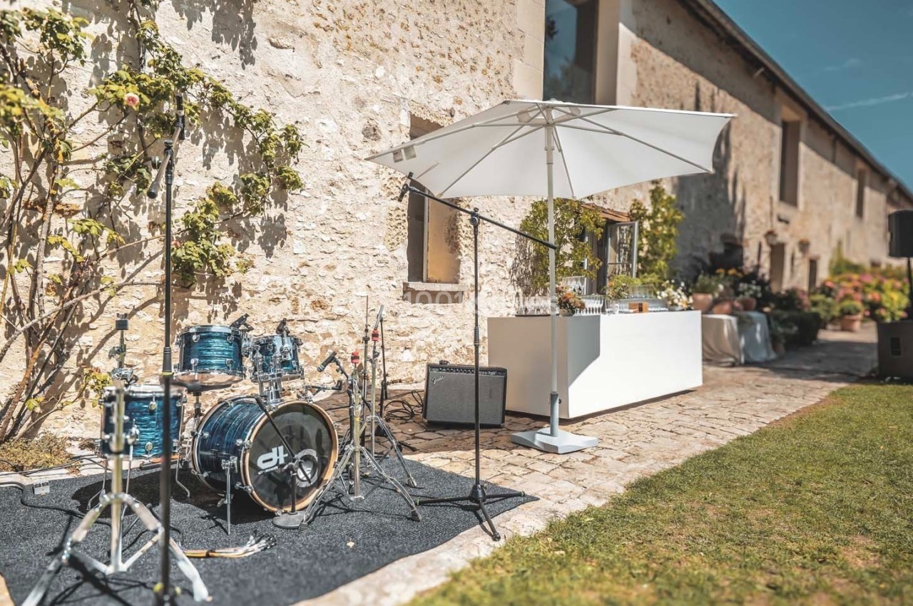 Batterie et micros installés en extérieur sur une terrasse pavée près d'un bar sous un parasol, devant un mur en pierre.