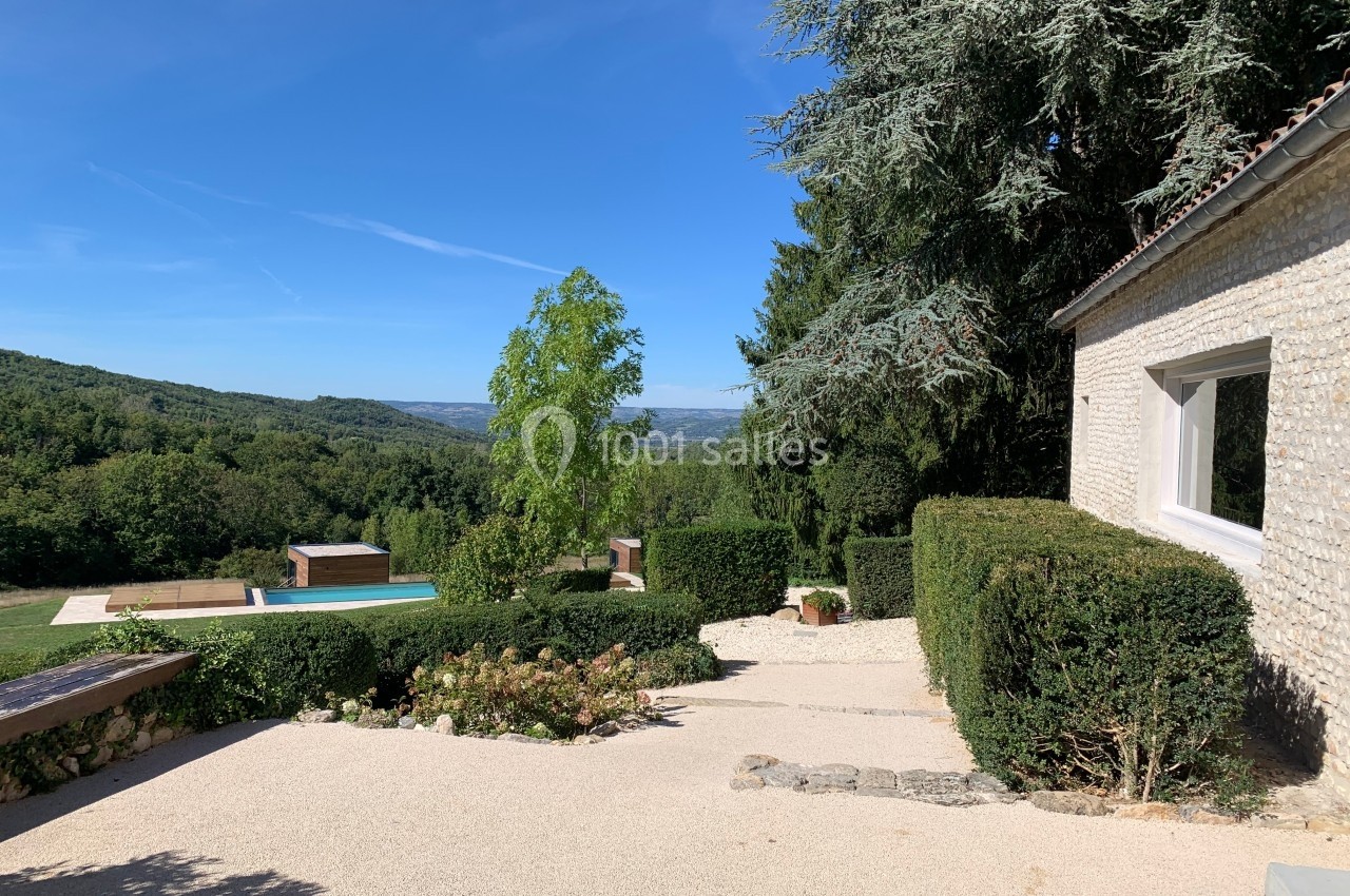 Vue d'une terrasse en gravier avec piscine, jardin verdoyant et collines sous un ciel bleu dégagé.