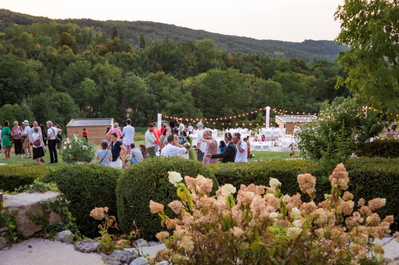 Groupe de personnes rassemblées dans un jardin verdoyant au crépuscule, avec des guirlandes lumineuses en arrière-plan.