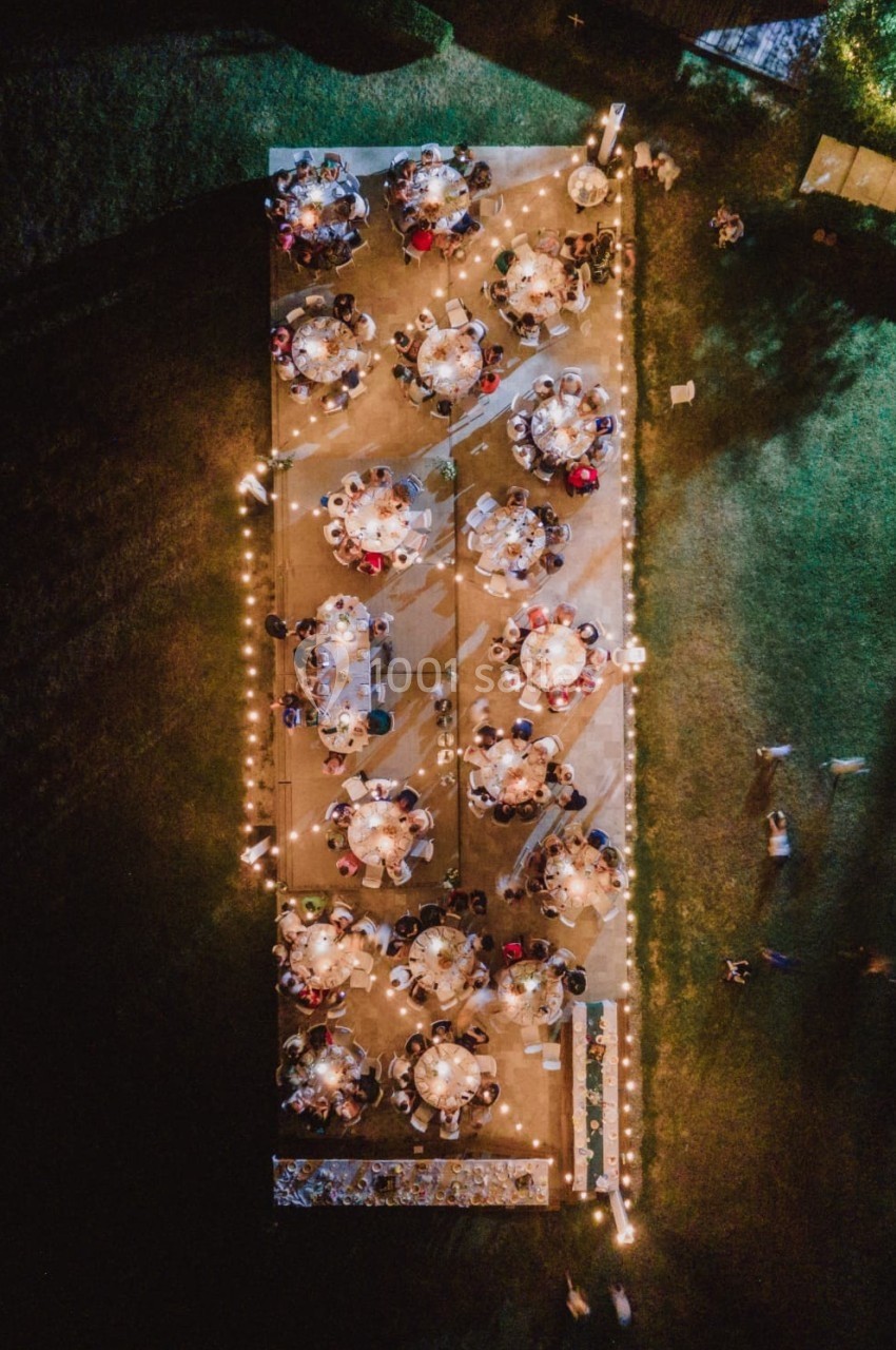 Vue aérienne d'un dîner en plein air avec des tables rondes éclairées par des guirlandes lumineuses dans un espace vert.