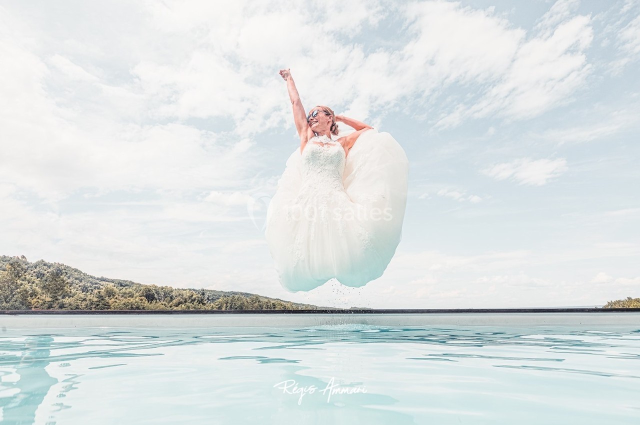 Une femme en robe de mariée saute au-dessus d'une piscine, ciel bleu et paysage en arrière-plan.
