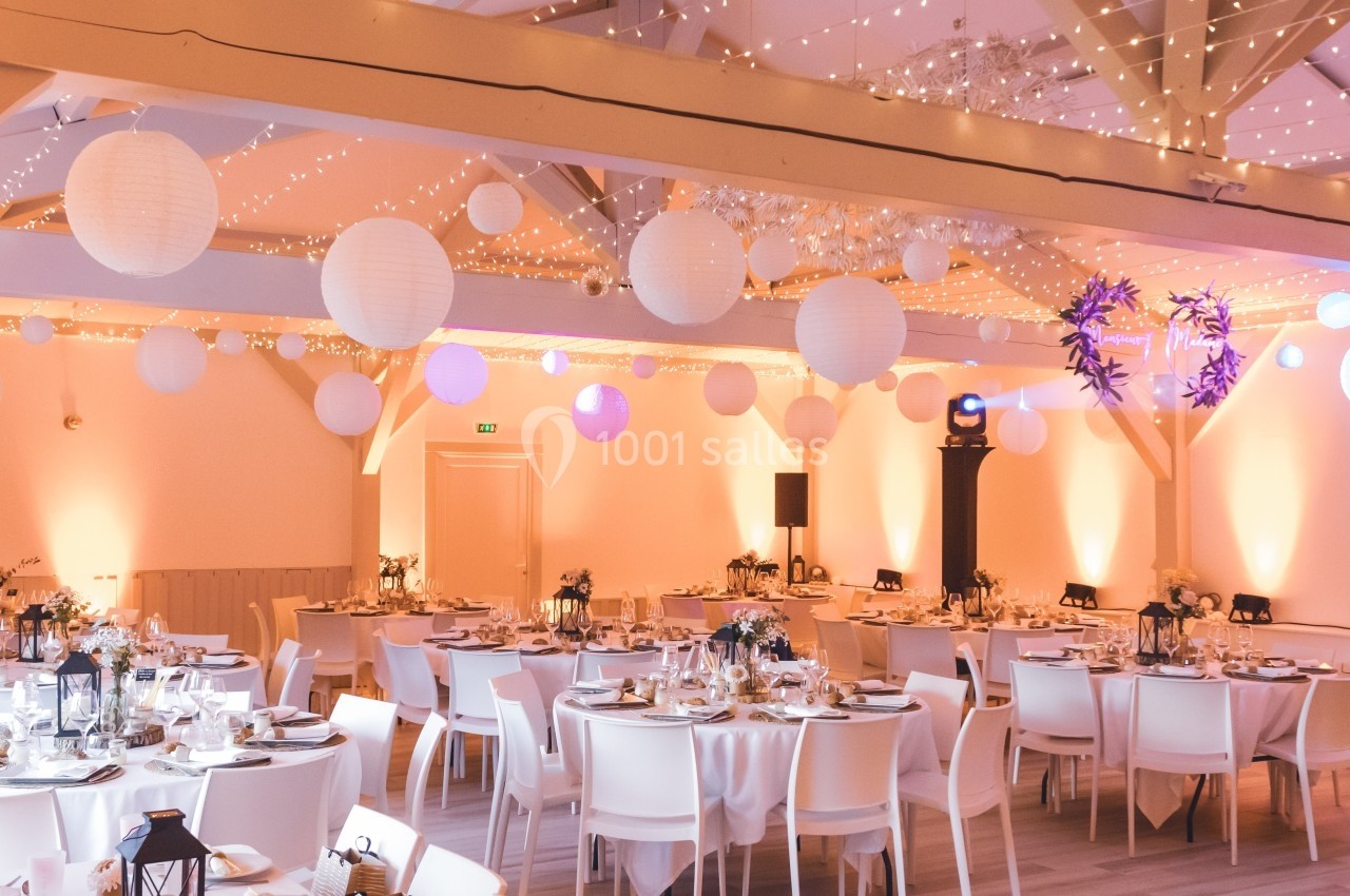 Salle de réception décorée avec des guirlandes lumineuses, des lanternes suspendues et des tables dressées pour un événement.
