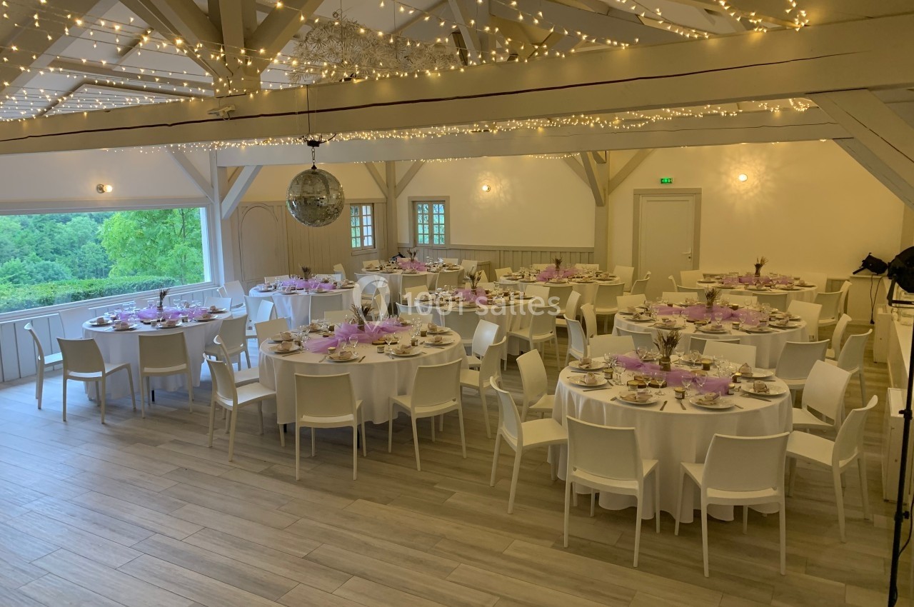 Salle de réception décorée avec des tables rondes dressées, guirlandes lumineuses suspendues et vue sur un paysage verdoyant.