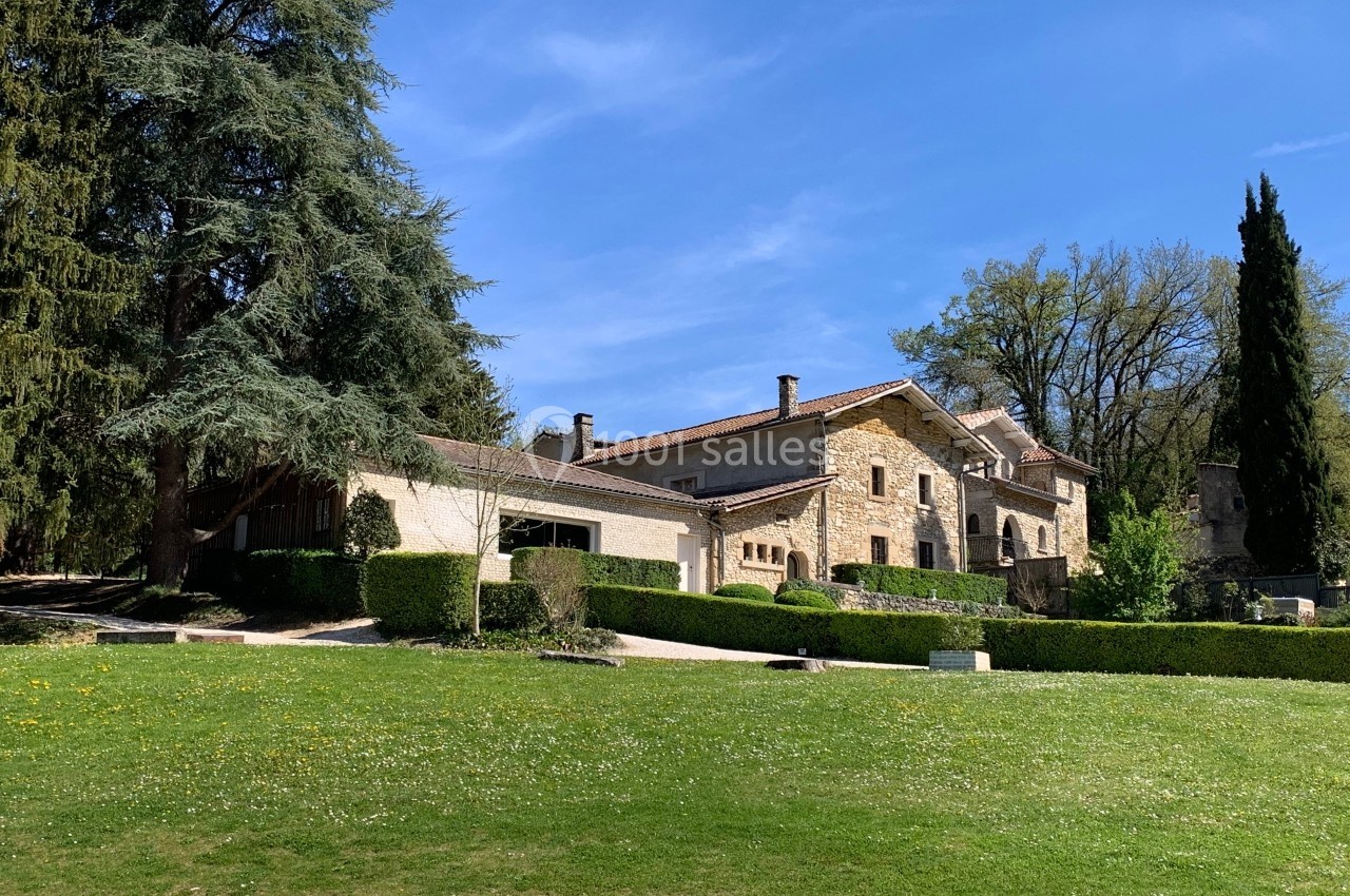 Grande maison en pierre entourée d'arbres et de pelouses, sous un ciel bleu dégagé.