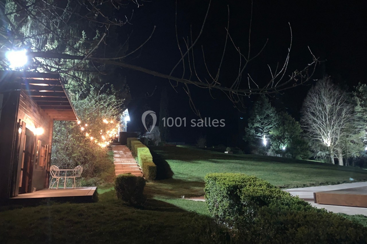 Jardin éclairé la nuit avec un chemin bordé de haies, une terrasse en bois et des guirlandes lumineuses.