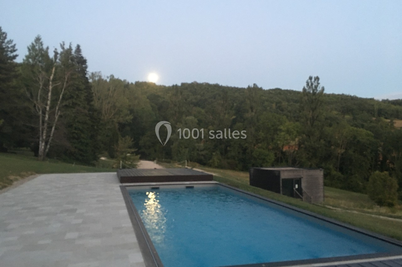 Piscine extérieure entourée de dalles, avec vue sur un paysage boisé et une pleine lune à l'horizon.