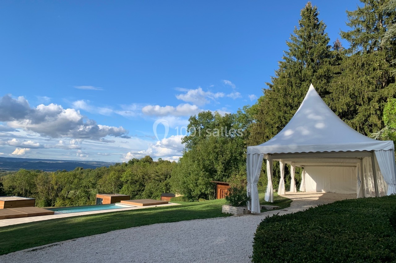 Tente blanche installée dans un jardin avec vue sur une piscine, des arbres et un paysage vallonné sous un ciel bleu.