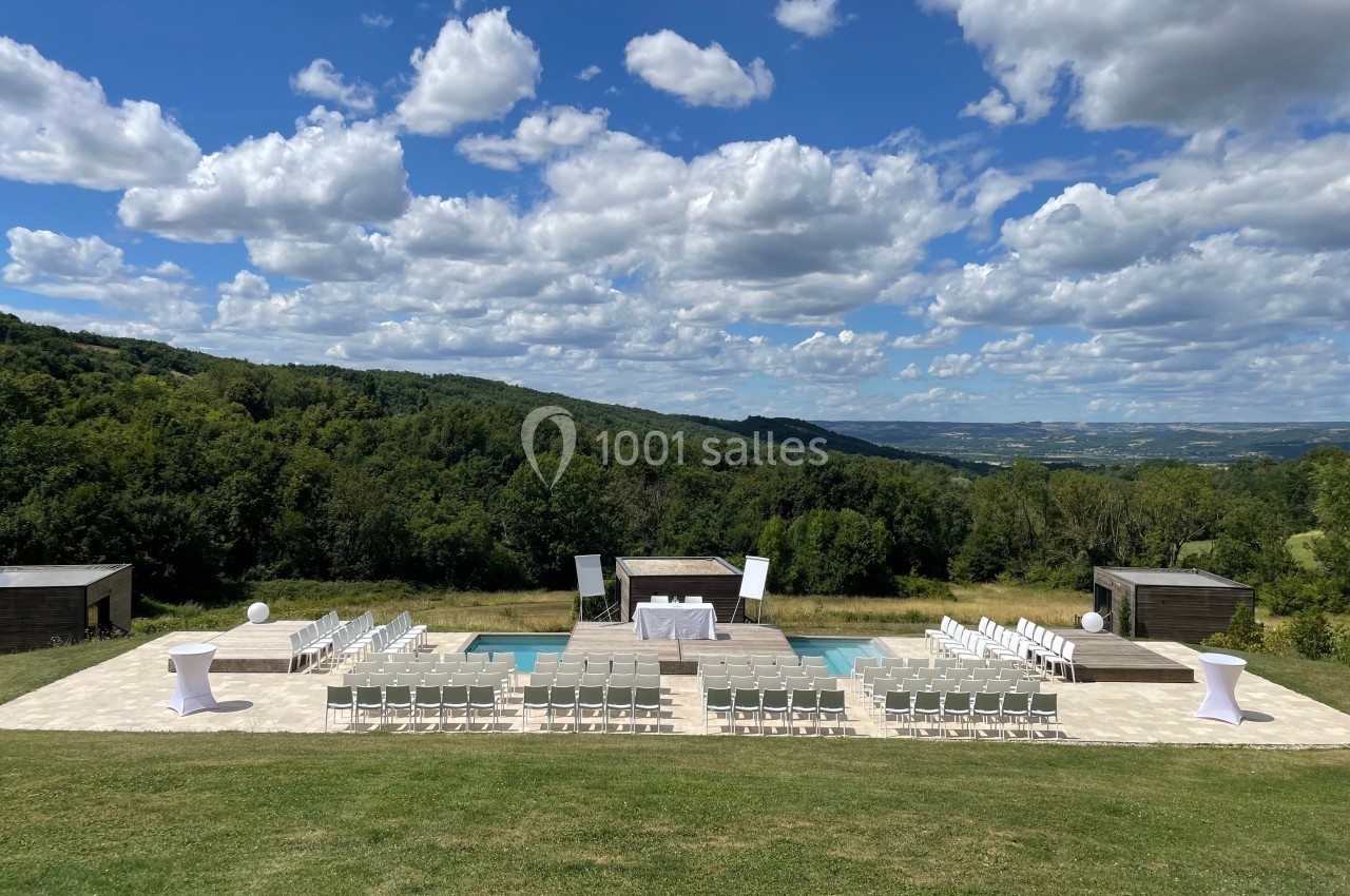 Chaises blanches disposées en extérieur pour une cérémonie, avec vue sur des collines verdoyantes sous un ciel partiellement…