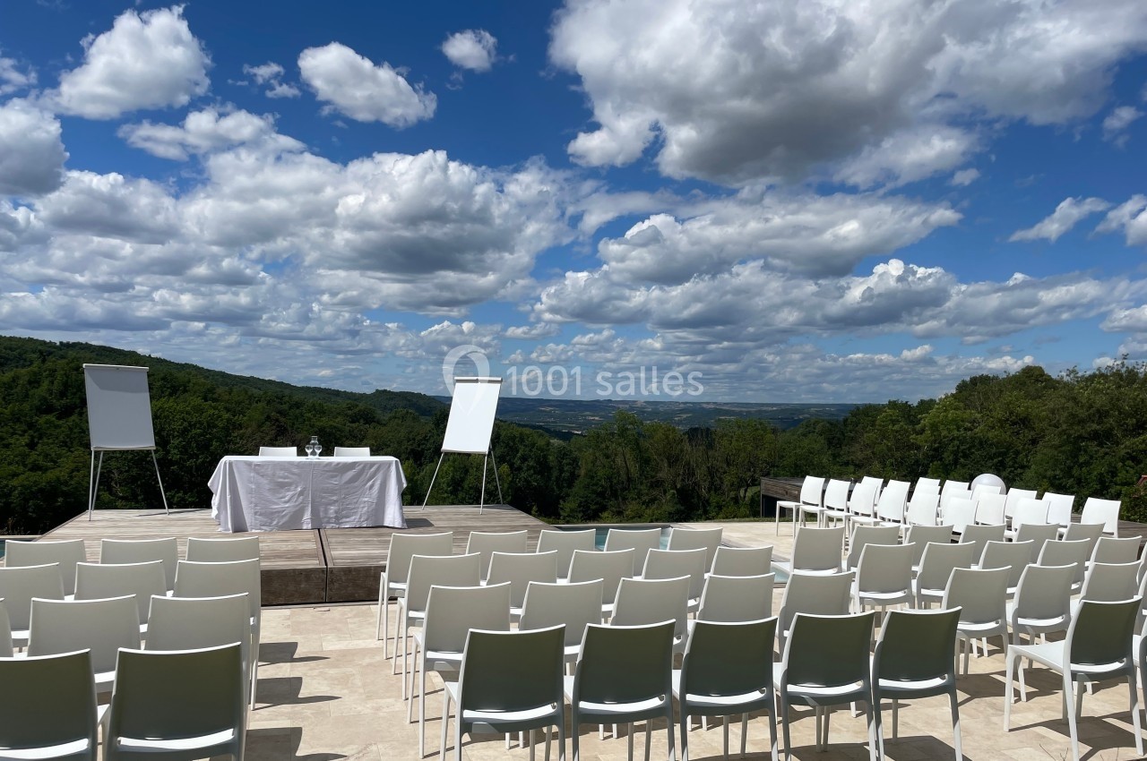 Chaises blanches alignées en extérieur face à une scène avec table, chevalets et vue sur un paysage vallonné.