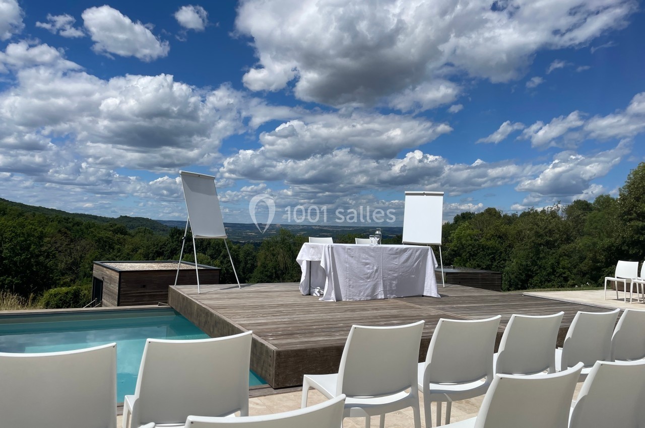 Plateforme en bois avec chevalets et table dressée, entourée de chaises blanches, vue sur un paysage verdoyant.
