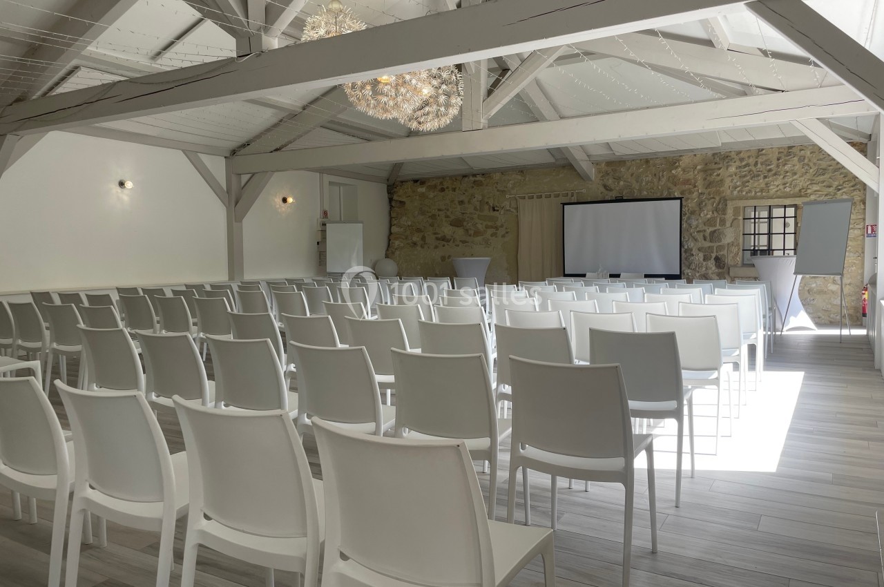 Salle de conférence lumineuse avec des rangées de chaises blanches, un écran de projection et un mur en pierre.