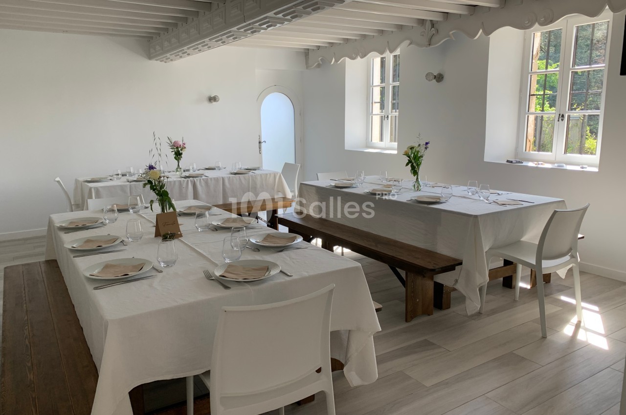 Salle lumineuse avec tables dressées, nappes blanches, chaises modernes et bancs en bois, près de fenêtres ouvertes.