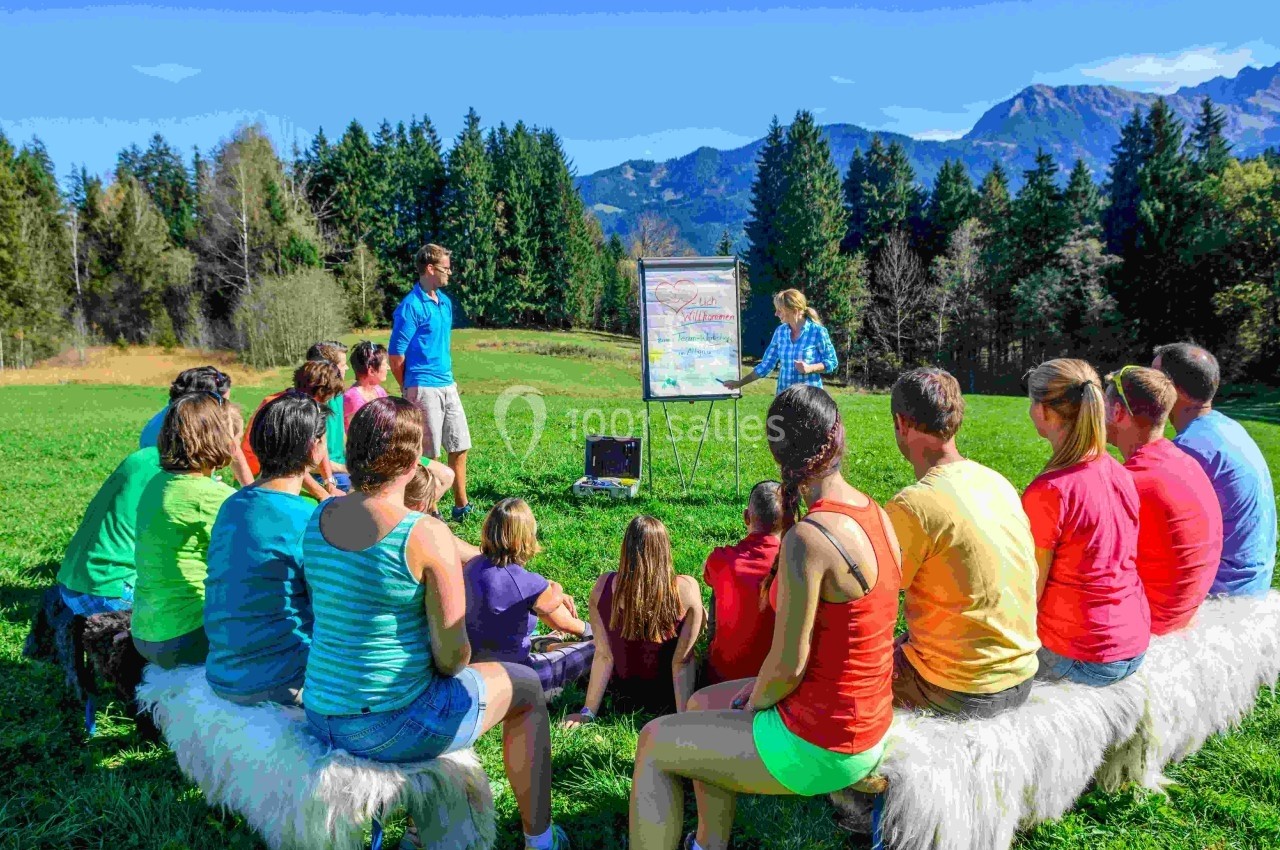 Un groupe de personnes assises en plein air écoute une présentation sur un tableau devant un paysage montagneux.