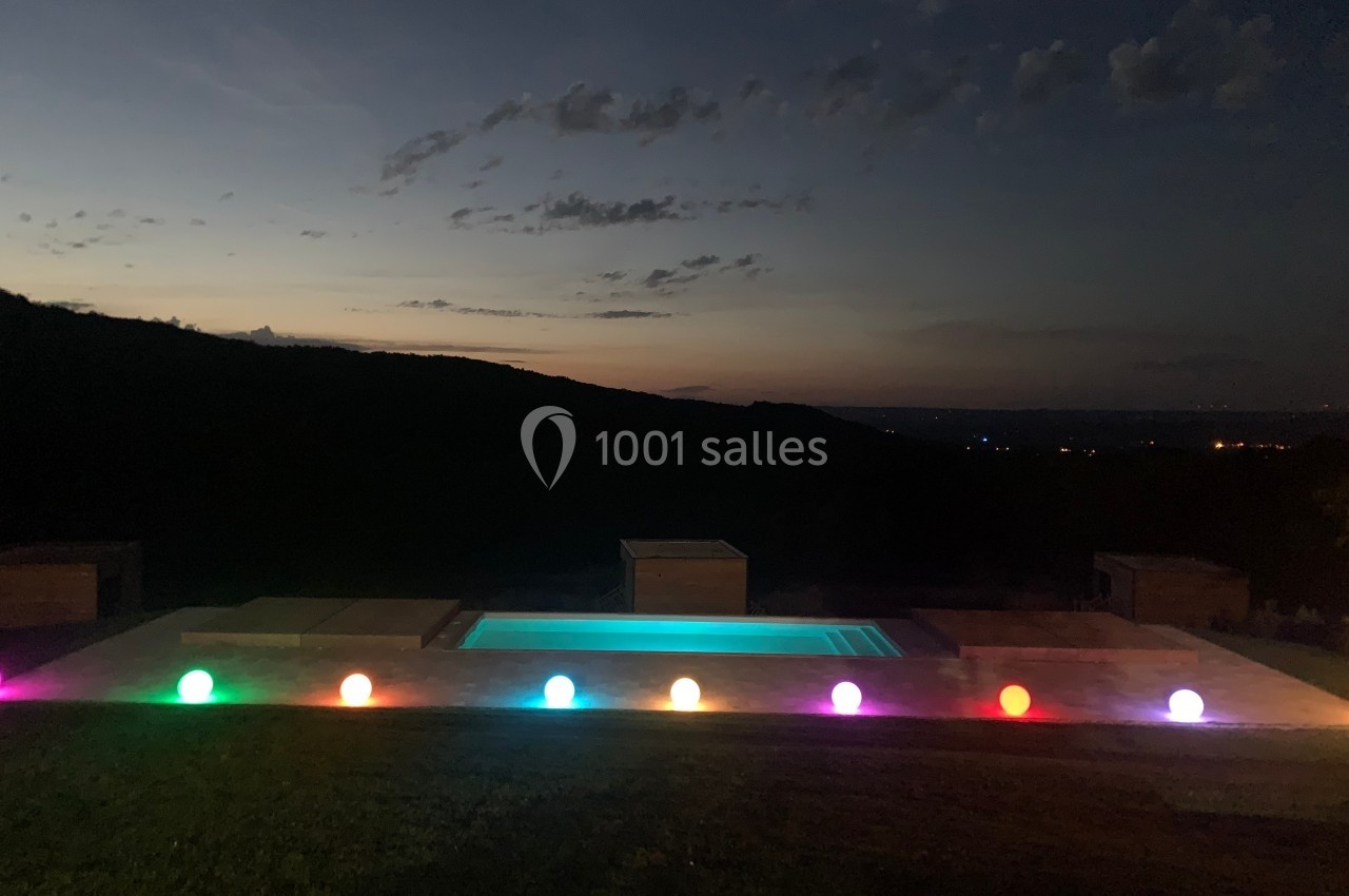 Piscine éclairée par des sphères lumineuses colorées, avec un paysage sombre et un ciel crépusculaire en arrière-plan.