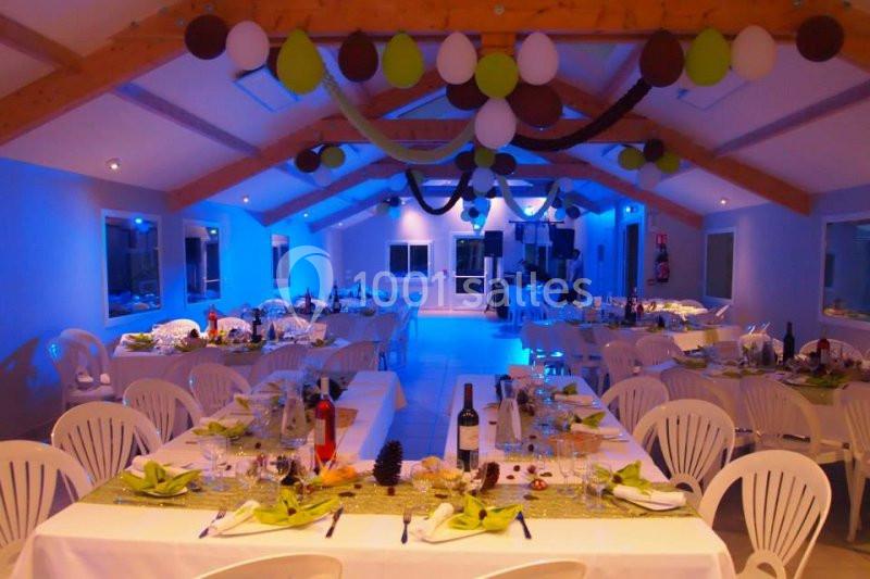 Salle décorée pour un événement avec tables dressées, nappes blanches, chaises en plastique et éclairage bleu.