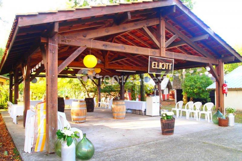 Abri en bois décoré de lanternes et guirlandes, avec tables, chaises blanches et tonneaux dans un cadre extérieur.