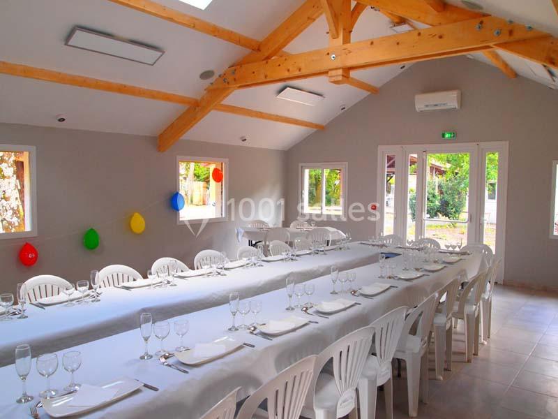 Salle lumineuse avec tables dressées, chaises blanches, poutres apparentes et décorations colorées accrochées aux murs.