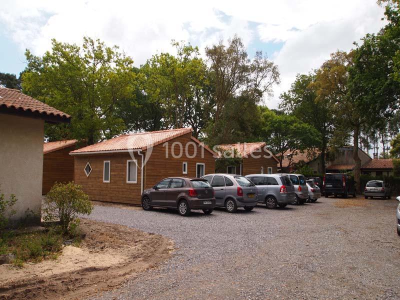 Alignement de voitures garées sur un parking en gravier, entouré de chalets en bois et d'arbres.