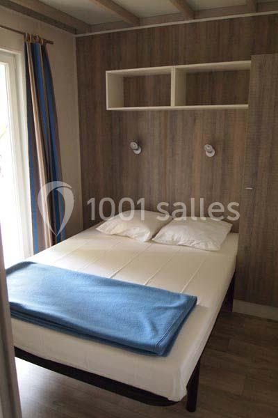 Lit double avec drap bleu, deux oreillers blancs, éclairage mural et étagères dans une chambre en bois clair.