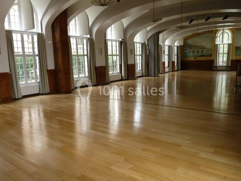 Salle spacieuse avec parquet en bois clair, grandes fenêtres cintrées et murs ornés de peintures murales.