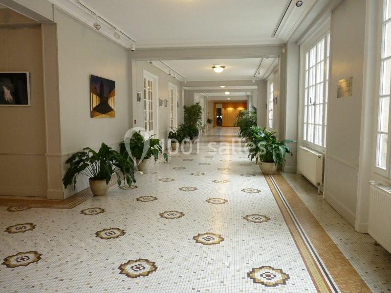 Couloir lumineux avec sol en mosaïque, plantes en pots et tableaux accrochés aux murs.