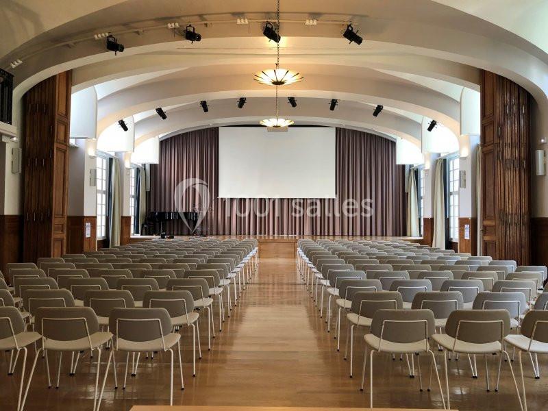 Salle de conférence vide avec des rangées de chaises blanches, une scène, un écran et des rideaux en arrière-plan.