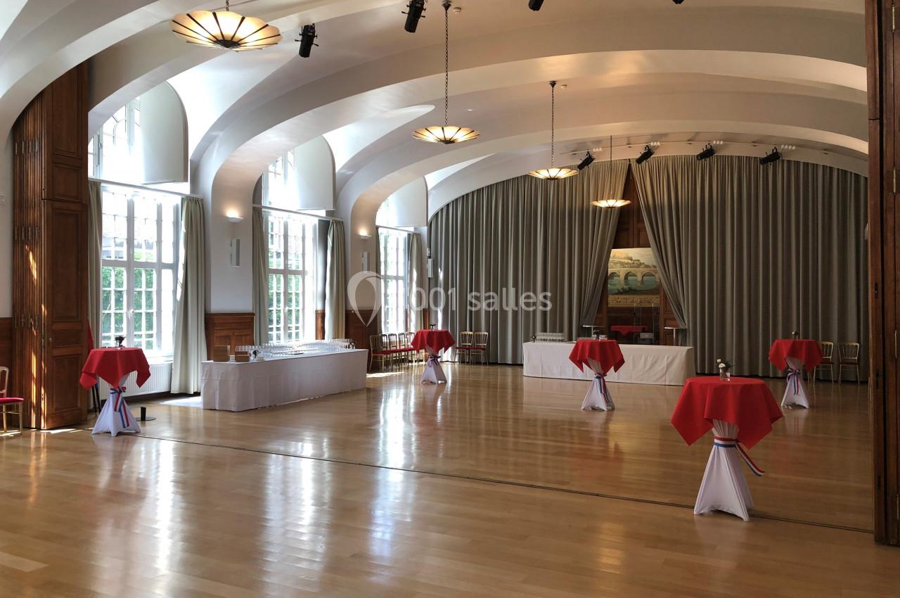 Salle de réception lumineuse avec parquet, tables hautes nappées de rouge et grandes fenêtres donnant sur l'extérieur.