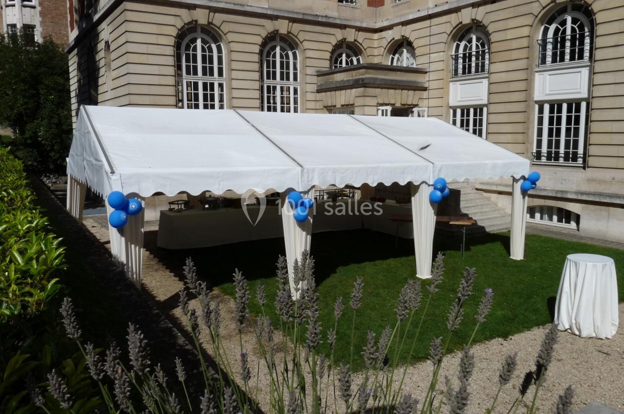 Tente blanche installée dans une cour avec des ballons bleus attachés aux poteaux et des tables en dessous.