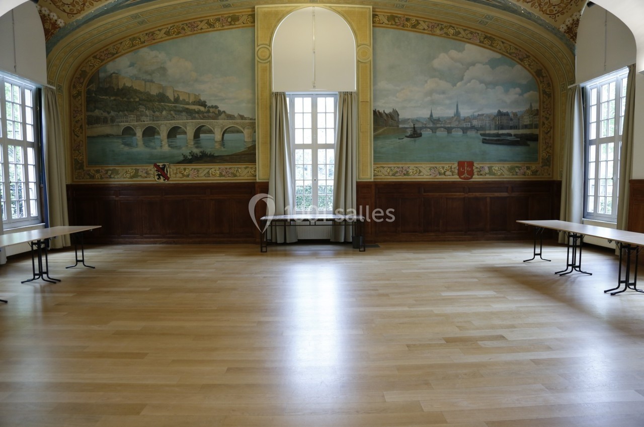 Salle vide avec parquet clair, murs ornés de fresques représentant un paysage fluvial et grandes fenêtres laissant entrer la…